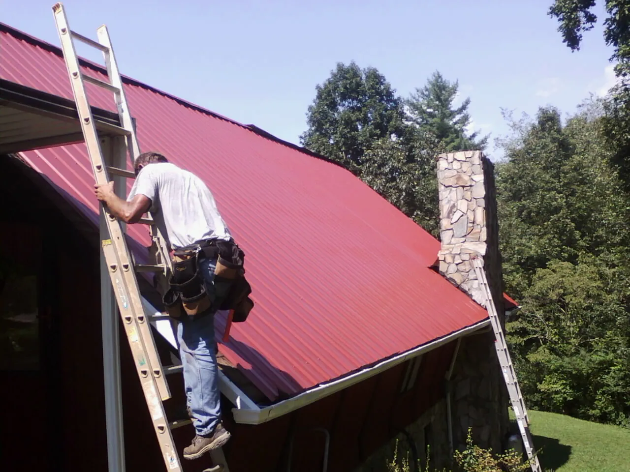 Roofer on a ladder beside a red metal roof and stone chimney — Soffit & Fascia Repair by Roofing Brothers in Mitchell